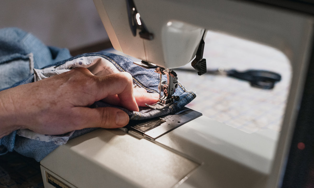 Close up of a person's hand pushing a set of denim jeans through a sewing machine.