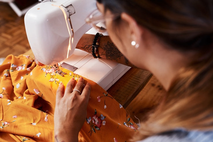 Woman sitting at a sewing machine stitching the edge of a piece of fabric.
